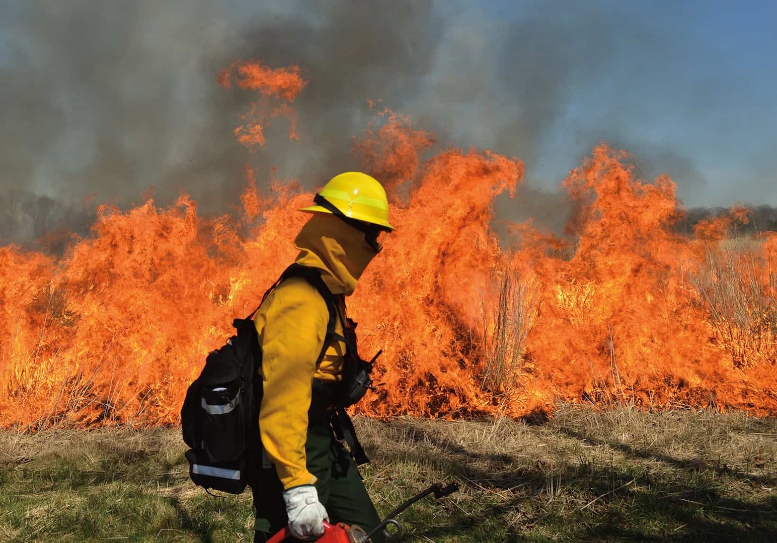 A person in fire safety gear walks in the foreground with orange flames licking at a meadow behind them.