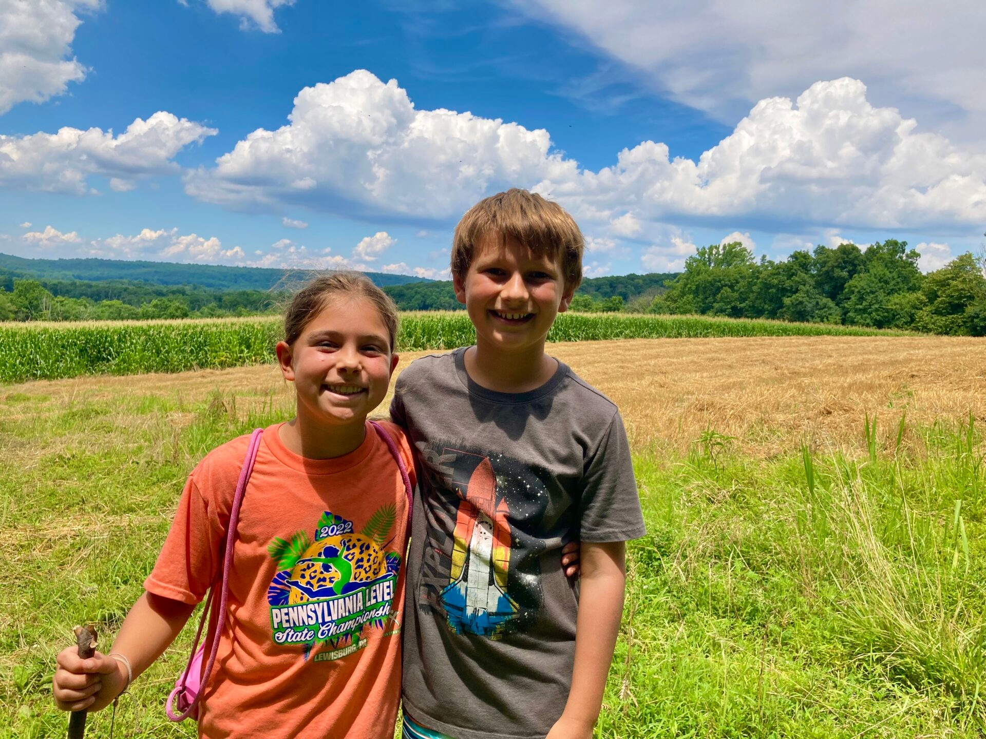 a young boy and girl standing with arms around each other smiling, one with a walking stick, with an agricultural field and blue sky with fluffy white clouds in the background