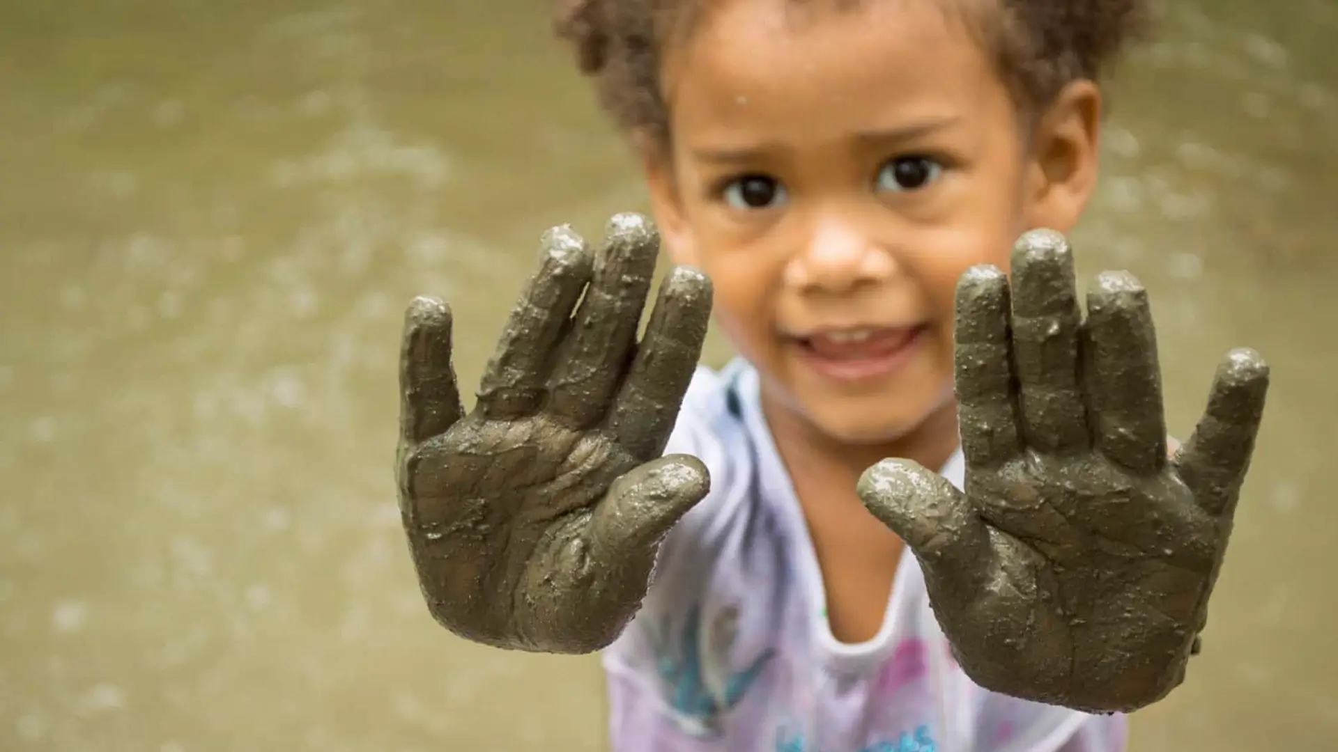 small girl stands in creek and holds out muddy hands to camera