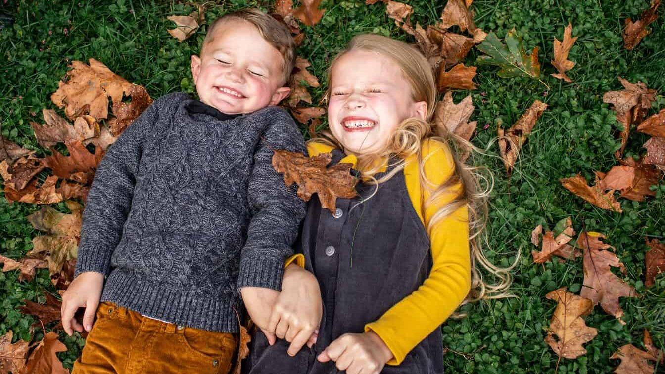 A girl and a boy in fall clothing smile with their eyes closed while lying on green grass and brown autumn leaves.