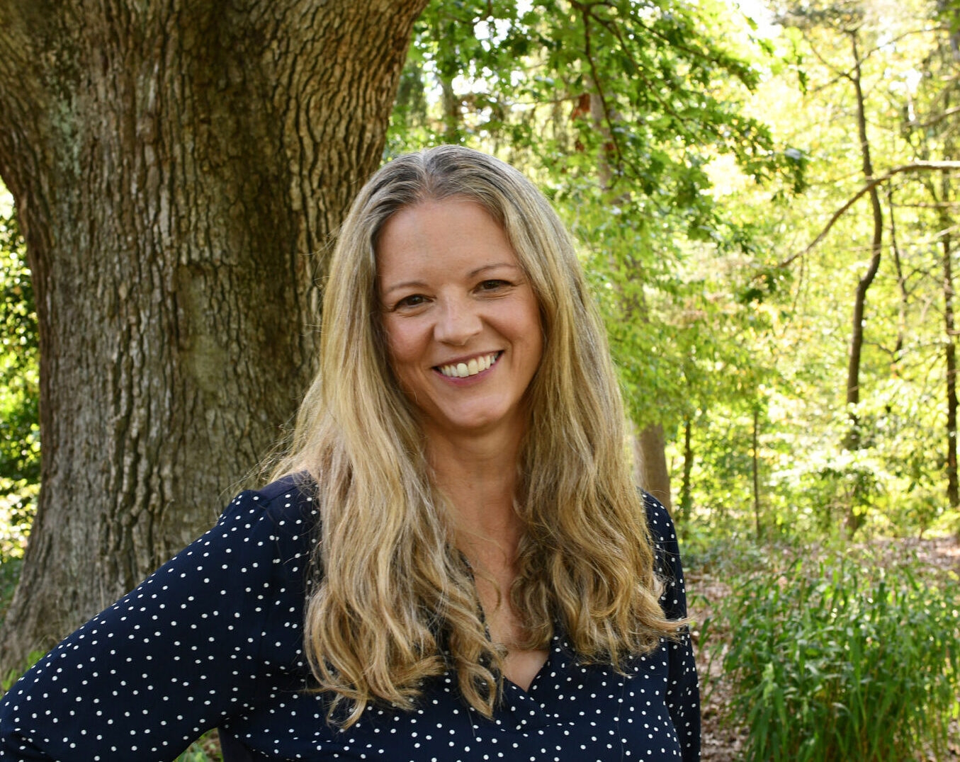 a smiling woman with long hair looking at the camera with a tree and forest in the background