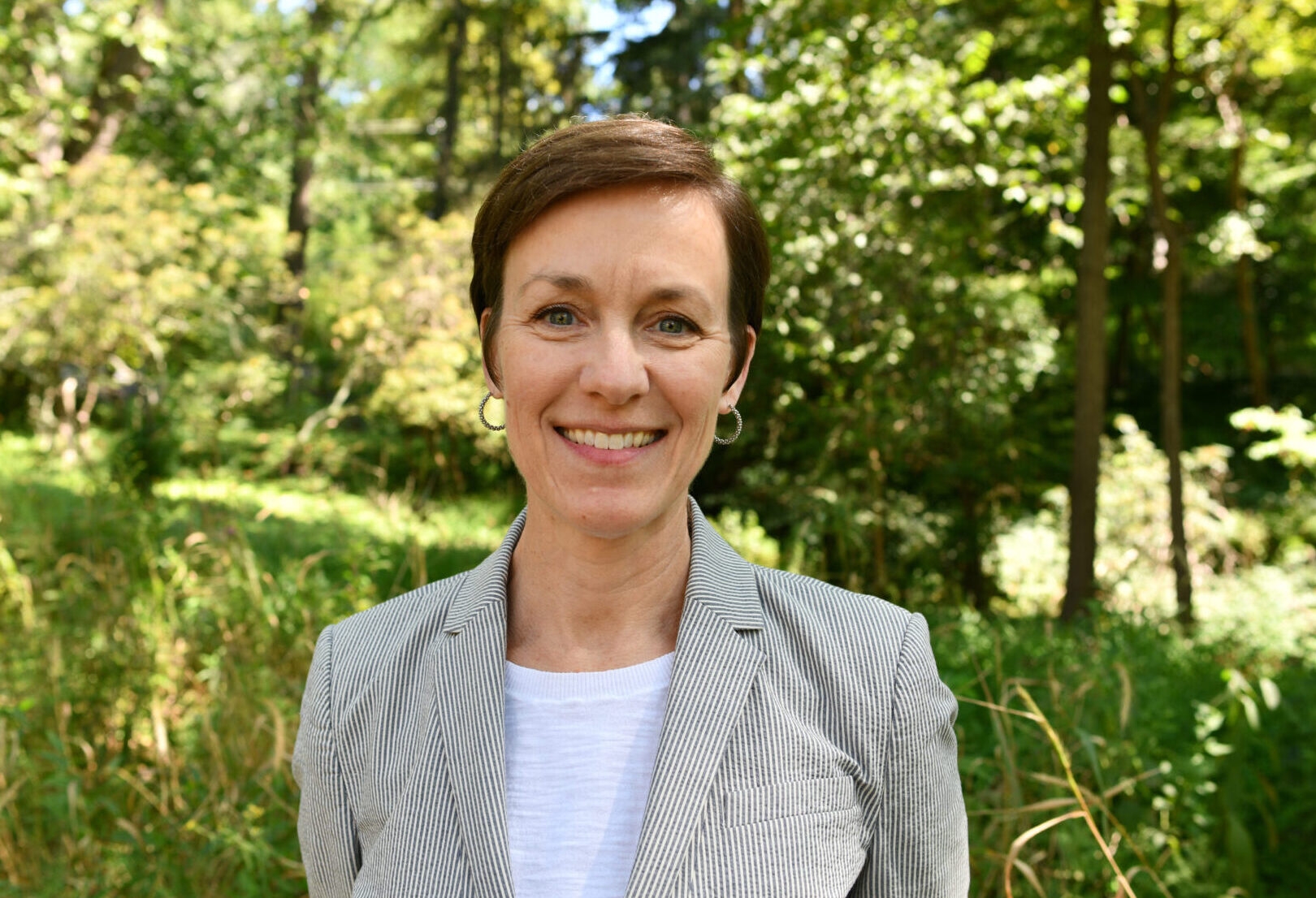 a woman wearing a striped blazer and earrings smiling at the camera standing in a forest