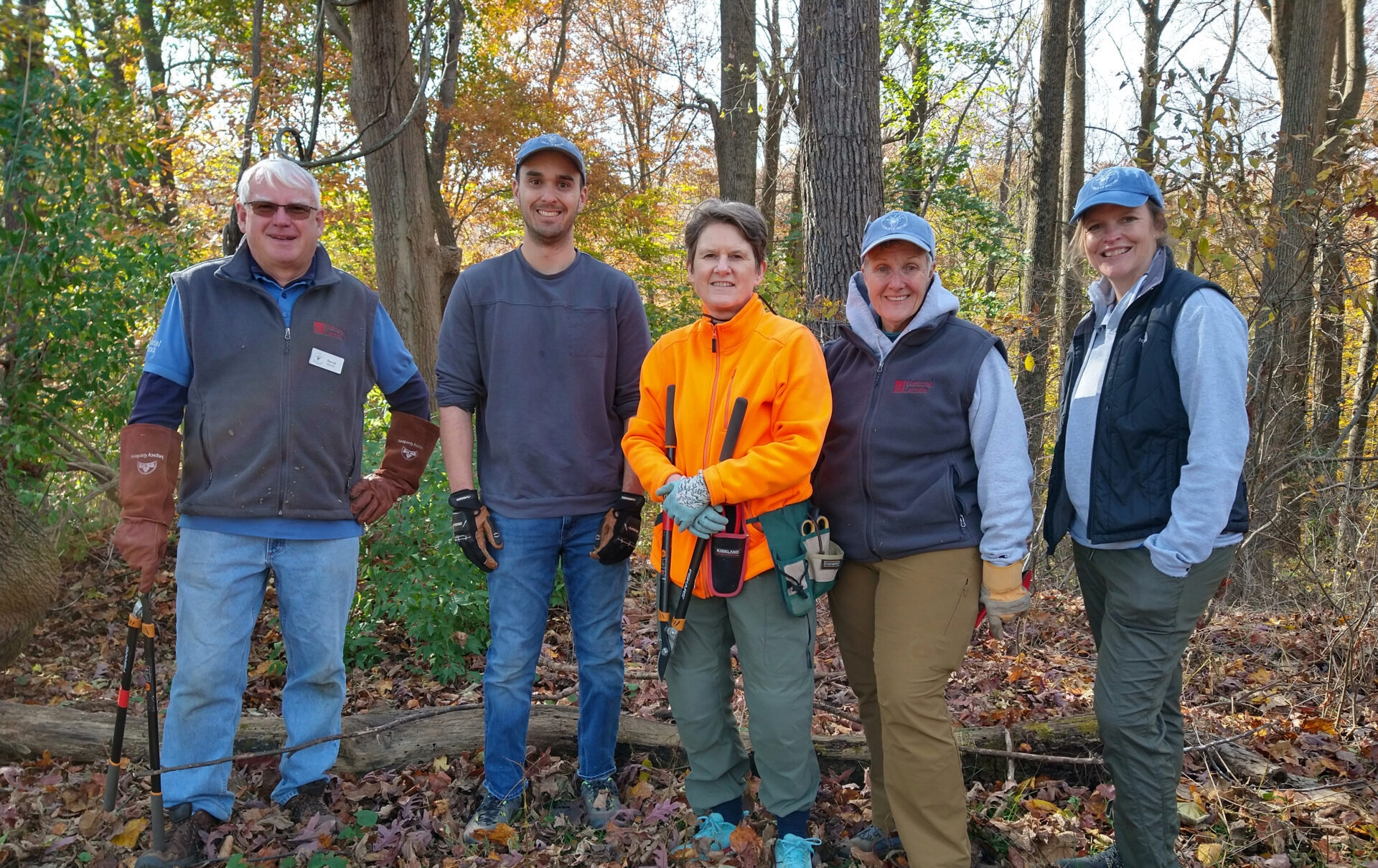 two men and three women smiling and holding garden tools in a forest with autumn leaves